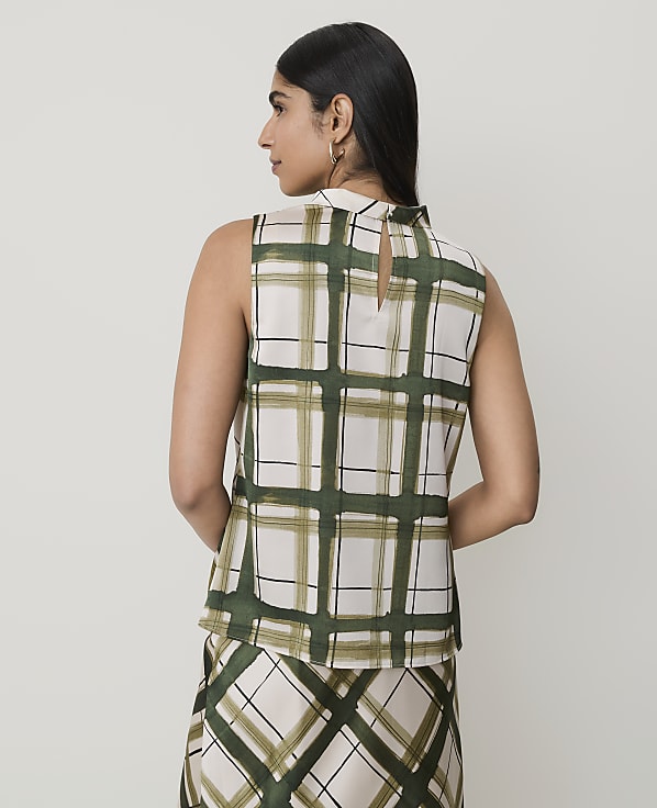 A woman in a sleeveless, high-neck, plaid top and matching skirt stands against a plain, light-colored background.