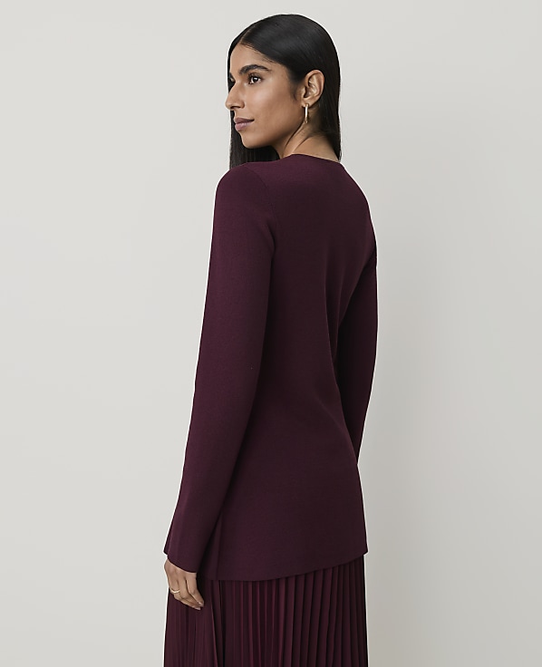 A woman in a burgundy sweater and pleated skirt stands against a plain, light-colored background.