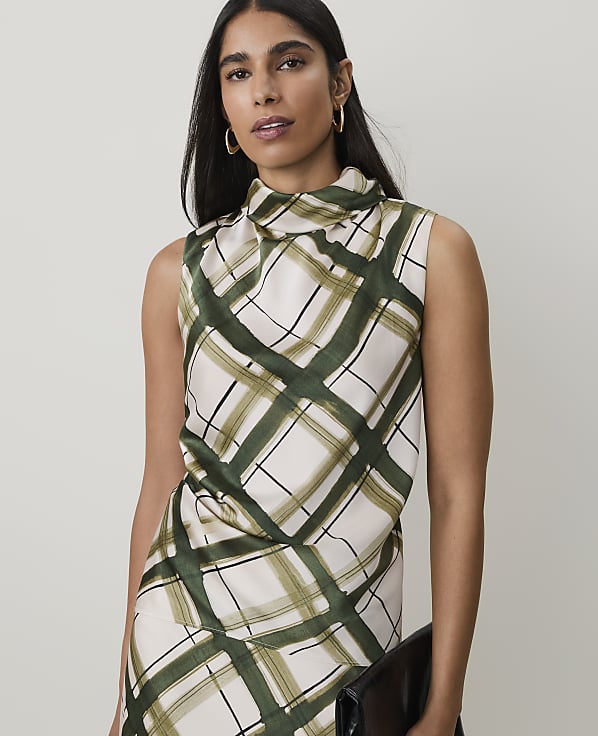 A woman in a cream and green plaid sleeveless top and holding a black clutch stands against a plain, light background.