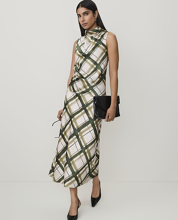A woman in a plaid dress and black heels stands against a plain background, holding a black clutch and sunglasses.