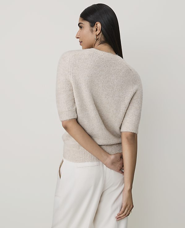 A woman in a cream sweater and pants stands against a plain, light background.