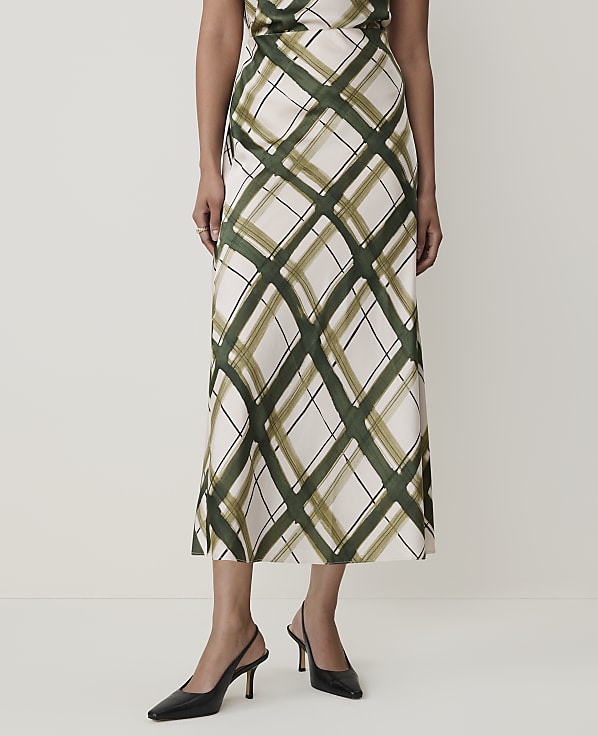 A woman in a cream and green plaid skirt and black slingback heels stands against a plain white background.