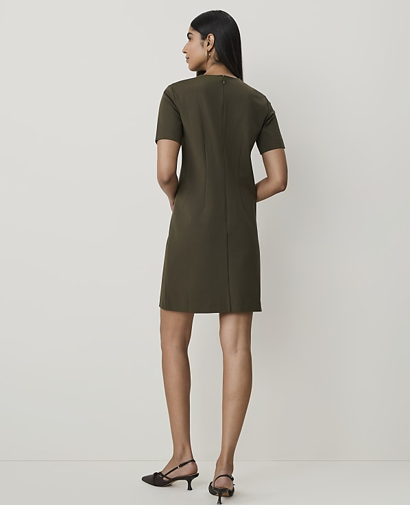 A woman in a short-sleeved olive green dress stands against a plain, light-colored background.
