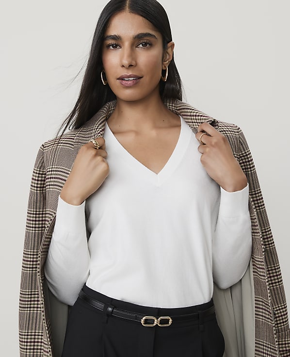 A woman in a white v-neck sweater and plaid blazer poses against a plain background.