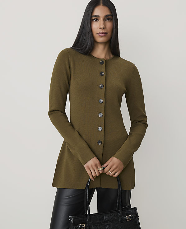 A woman in a green cardigan and black pants holds a black purse in front of a plain background.