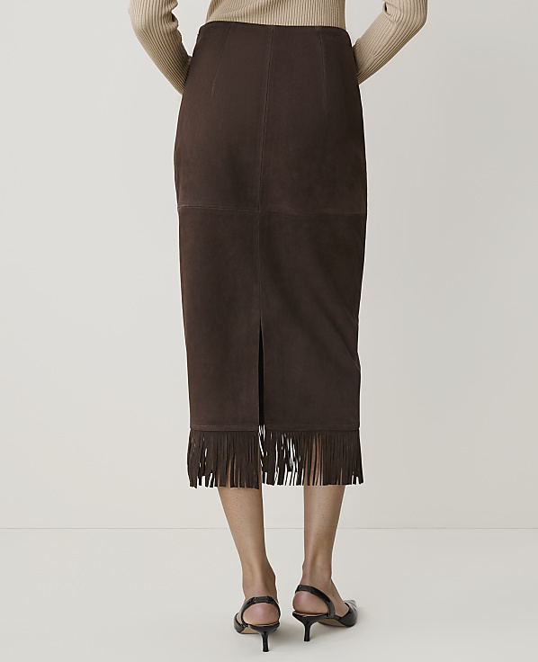 A person stands in front of a plain background, wearing a brown suede skirt with fringe at the hem and black heeled shoes.