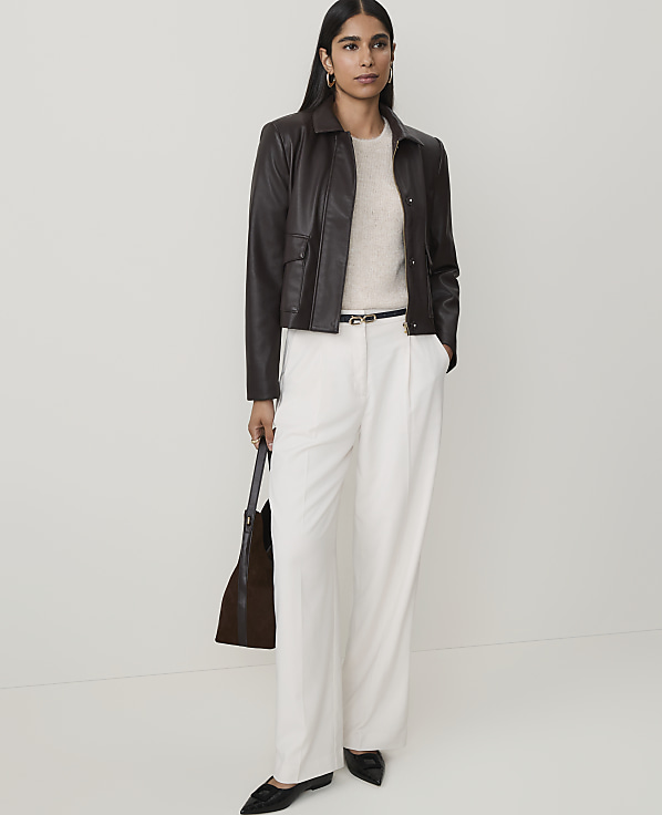 A woman in a cream sweater, dark brown leather jacket, and white wide-leg pants stands against a plain white background, holding a dark brown tote bag.