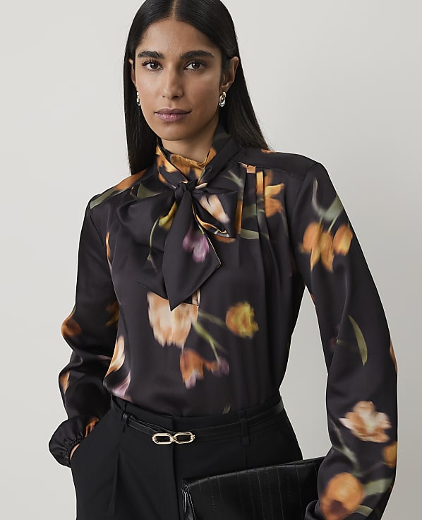 A woman in a floral print blouse and black pants holds a clutch in front of a plain background.