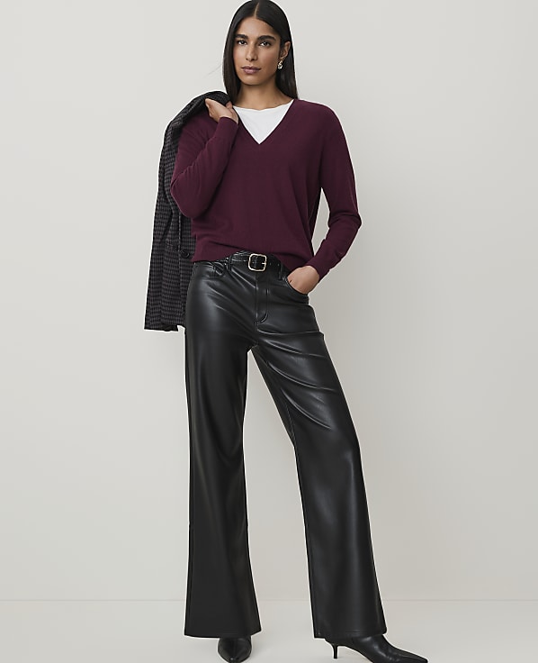 A woman in a burgundy v-neck sweater and black leather pants stands against a plain, light gray background, with a blazer draped over her shoulder.