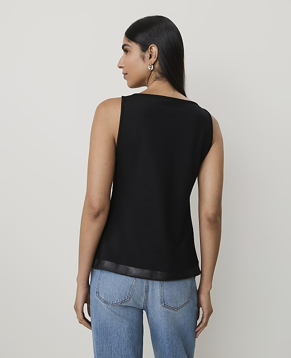 A woman in a black sleeveless top and blue jeans stands against a plain, light-colored background.