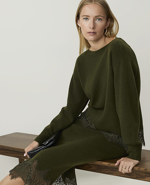 A woman in a green sweater and skirt sits on a wooden bench against a plain background.