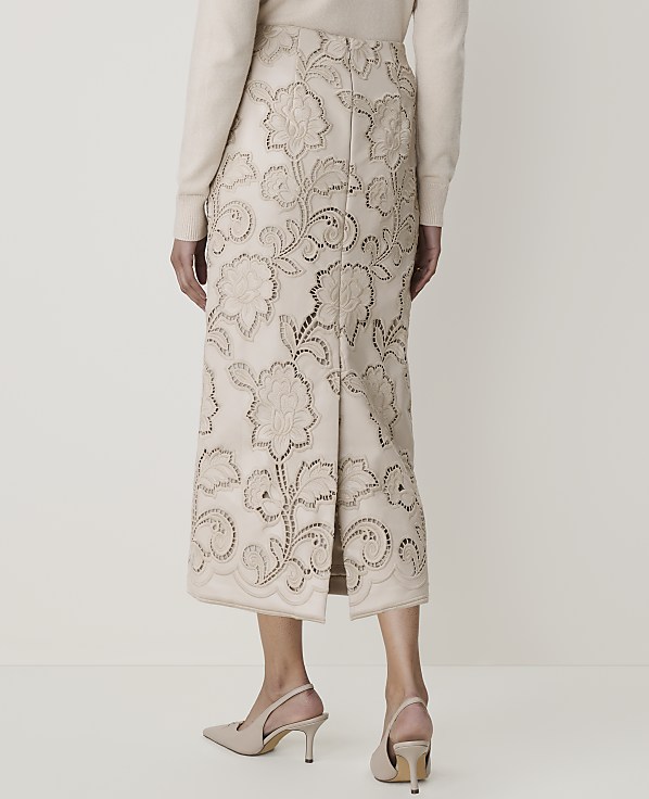 A woman in a cream sweater and a cream embroidered skirt with floral cutouts stands against a plain cream background.