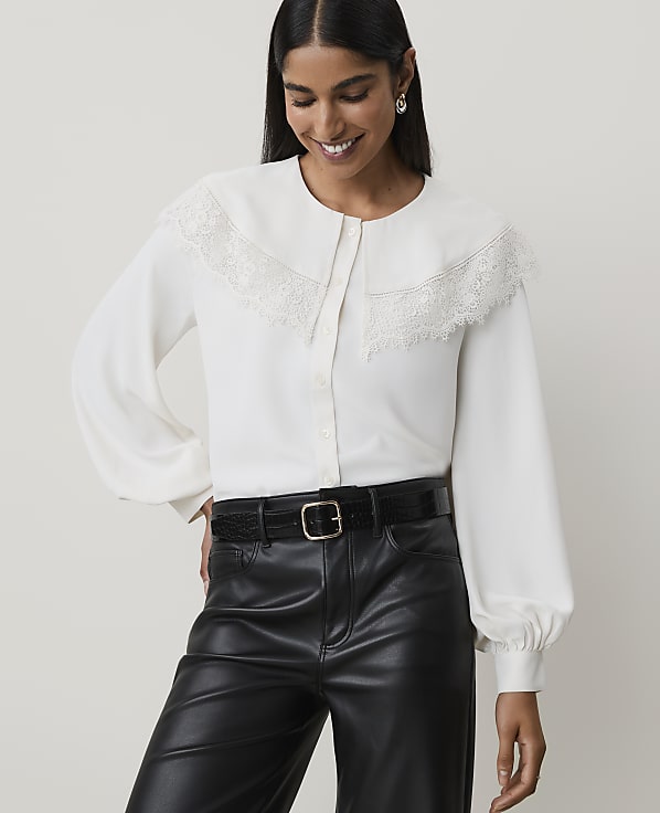A woman in a white lace-trimmed blouse and black leather pants smiles against a plain background.