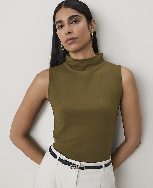 A woman in a sleeveless olive green turtleneck stands against a plain, light gray background, wearing white pants and a dark belt.