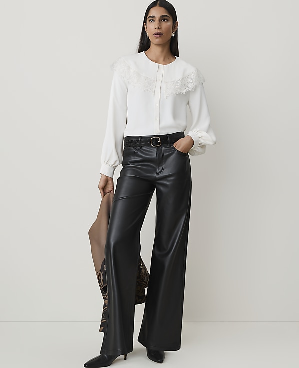 A woman in a white blouse and black leather pants stands against a plain white background, holding a brown jacket.