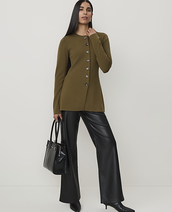 A woman in a green asymmetrical jacket and black leather pants stands with a black handbag against a plain background.