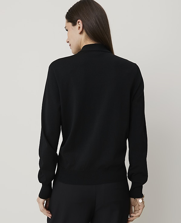 A woman in a black turtleneck sweater and pants stands against a plain white background.