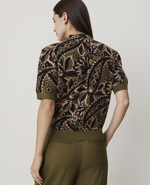 A woman in a patterned top and olive pants stands against a plain background.