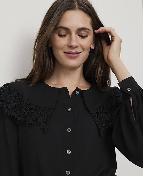 A woman with long brown hair smiles gently, her hand touching her hair, while wearing a black blouse with a large lace collar against a plain white background.