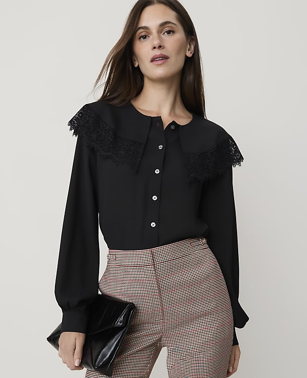 A woman in a black blouse and plaid pants holds a black clutch in front of a plain background.
