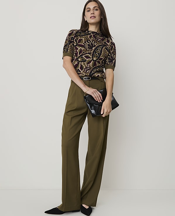 A woman in a patterned top and olive green pants holds a black clutch in front of a plain, light-colored background.