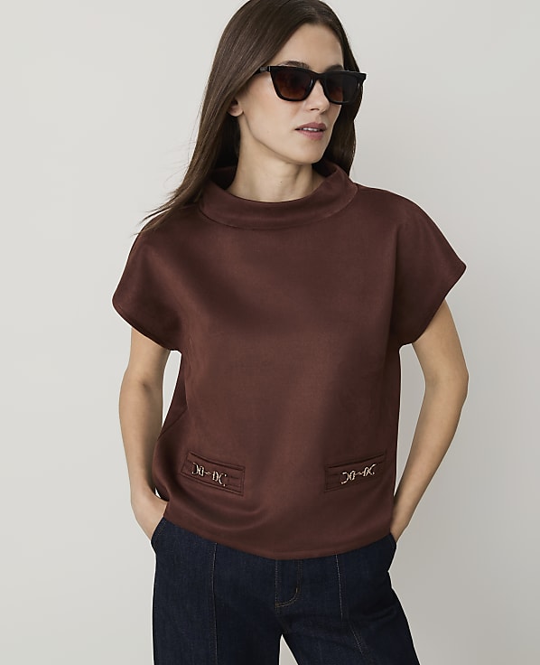 A woman in a brown top and dark jeans stands against a plain white background.