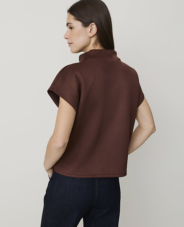 A woman in a brown mock-neck top and dark jeans stands against a plain, light gray background.