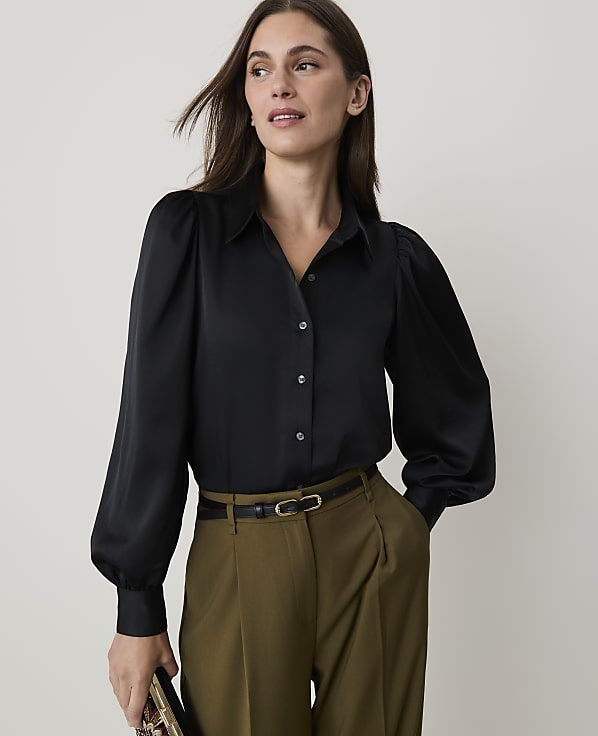 A woman in a black satin blouse and olive green pants stands against a plain, light background.
