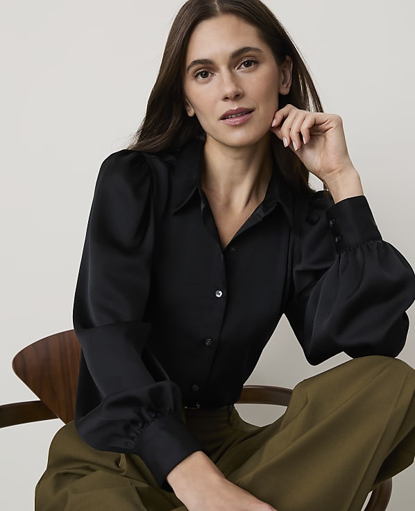 A woman in a black satin blouse and olive green pants sits on a wooden chair against a plain background.