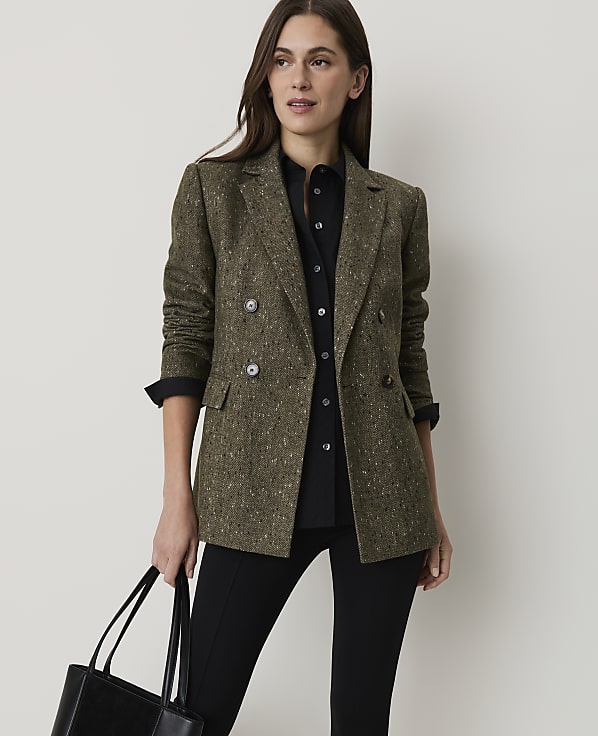 A woman in a tweed blazer and black shirt holds a tote bag in front of a plain background.