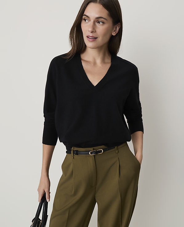 A woman in a black v-neck sweater and olive green pants stands against a plain, light-colored background.