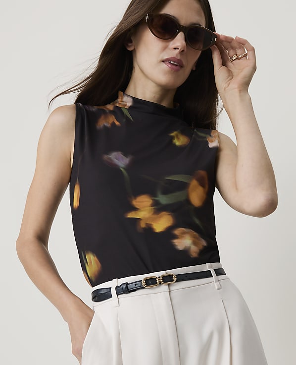 A woman in a floral top and light-colored pants adjusts her sunglasses against a plain background.