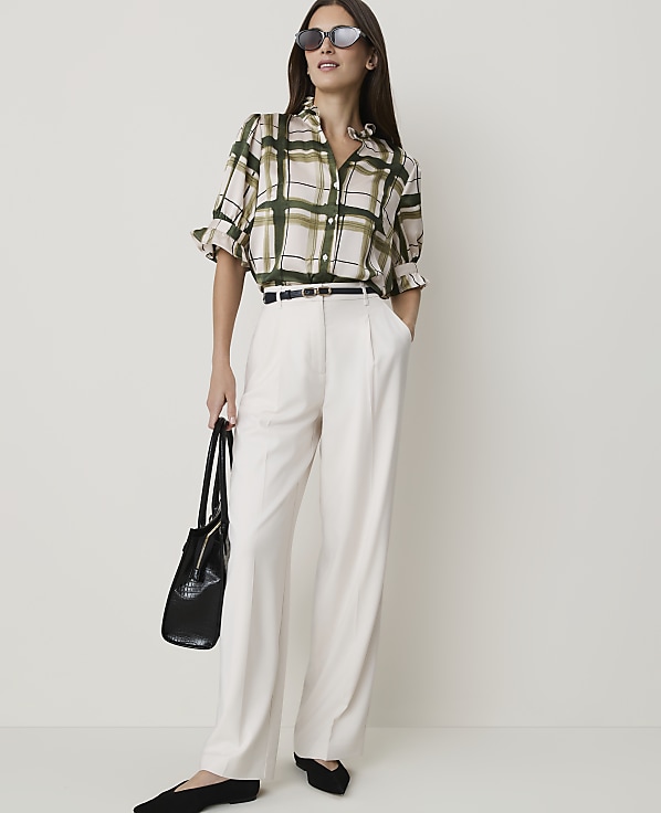 A woman in a plaid blouse and cream pants stands against a plain white background, holding a black handbag.