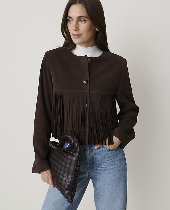 A woman in a brown suede jacket with fringe and blue jeans holds a dark woven bag in front of a plain white background.