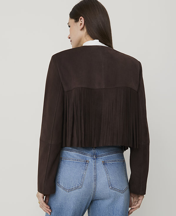 A woman in blue jeans and a brown suede jacket with fringe stands against a plain white background.