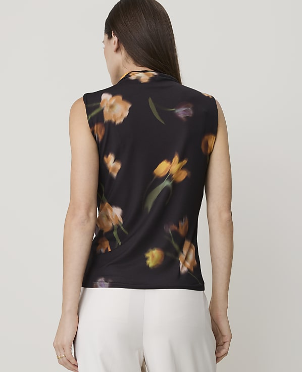 A woman in a black floral-print sleeveless top stands against a blurred background.