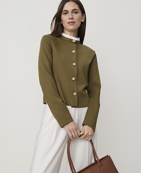 A woman in a green cardigan and white pants holds a brown tote bag in front of a plain background.