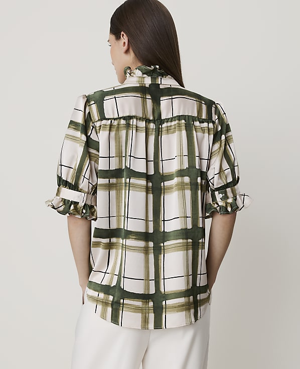 A woman in a light pink and green plaid blouse stands against a plain white background.
