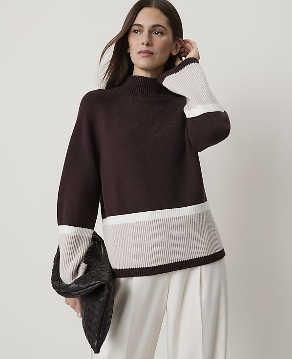 A woman in a burgundy sweater with cream and beige stripes holds a dark clutch in front of a plain background.