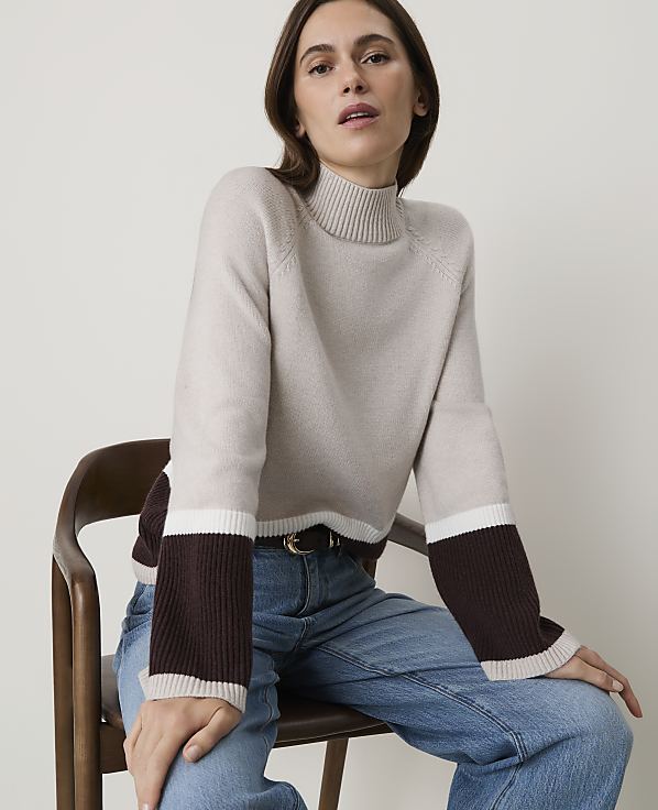 A woman in a color-blocked sweater and jeans sits on a wooden chair against a plain background.