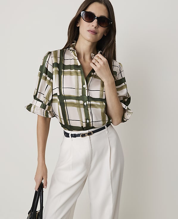 A woman in a plaid blouse and white pants stands against a plain background.