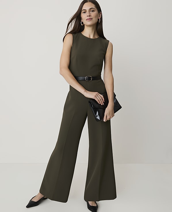 A woman in a green jumpsuit and black heels stands with a clutch in front of a plain white background.