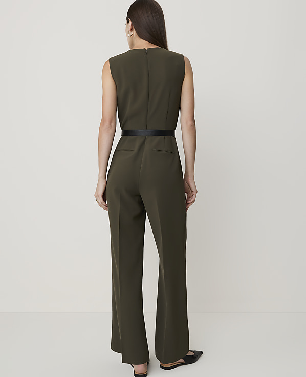 A woman in a sleeveless olive green jumpsuit with a black belt stands against a plain, light-colored background.