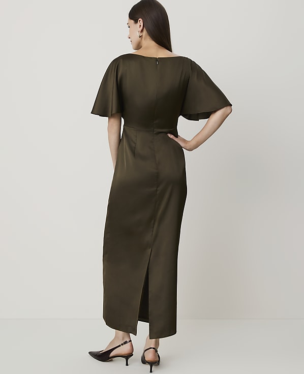 A woman in a dark olive green dress with flutter sleeves stands against a plain, light background.