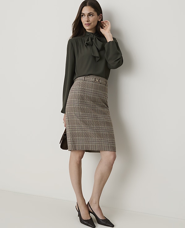 A woman in a dark green bow-neck blouse and a plaid pencil skirt stands against a plain, light-colored wall.