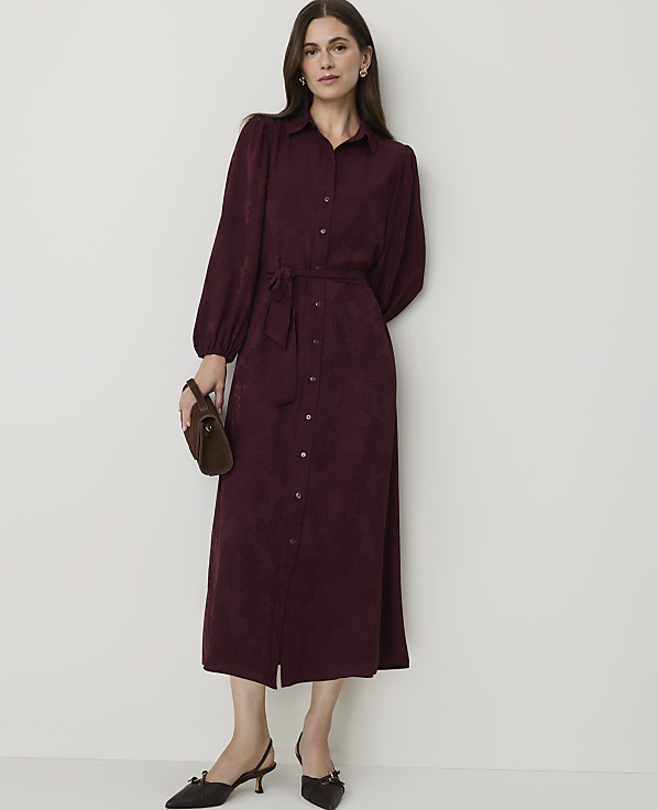 A woman in a burgundy shirtdress and pointed-toe heels stands against a plain, light-colored wall, holding a small clutch.