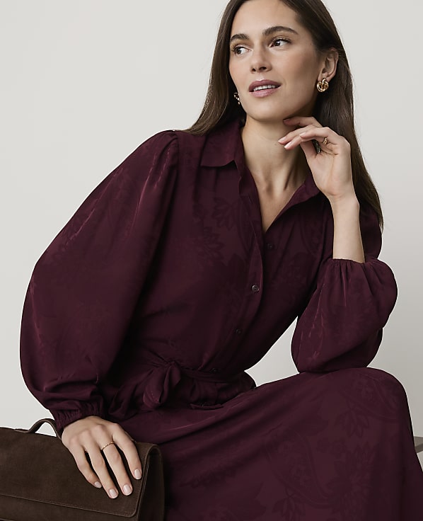 A woman in a burgundy dress with puffed sleeves poses against a plain, light-colored background, with a brown bag in the foreground.
