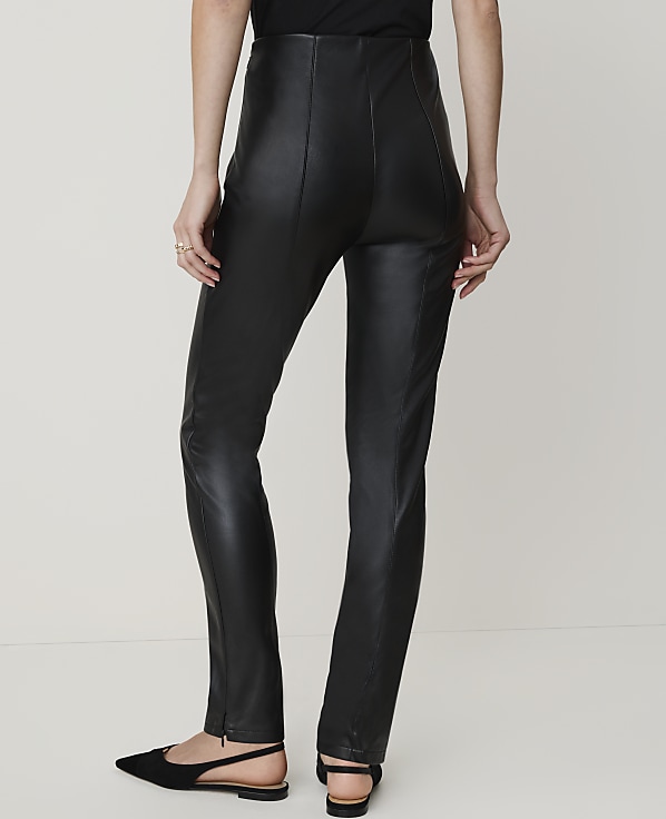 A person wearing black faux leather pants and black pointed-toe flats stands against a plain white background.