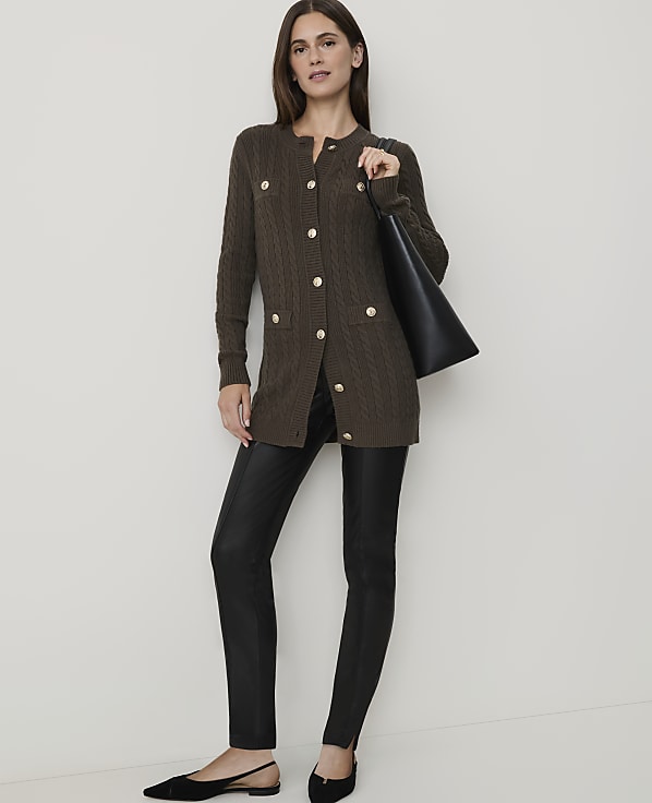 A woman in a brown cable-knit cardigan and black leather pants stands against a plain white background, holding a black tote bag.