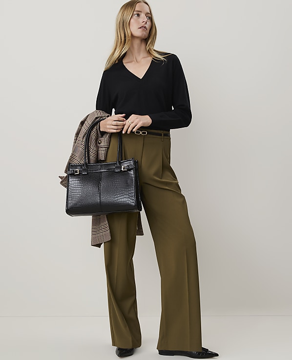 A woman in a black v-neck sweater and olive green wide-leg pants stands holding a black crocodile-embossed handbag and a plaid jacket over her shoulder against a plain white background.